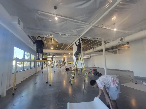 Interior of a large commercial space with plastic sheeting covering the floor and walls, scaffolding, and exposed insulation in the process of being installed.