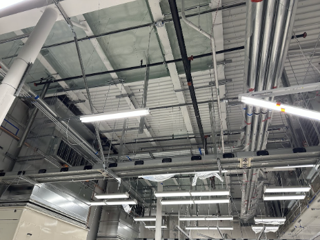 A ceiling view of an industrial building showing exposed pipes, ductwork, and fluorescent lighting fixtures.
                     