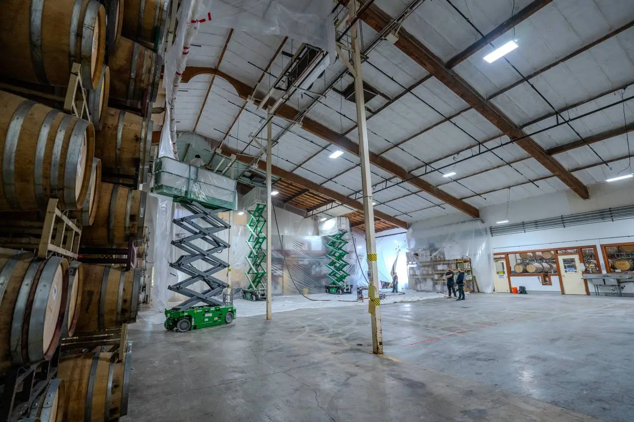 Interior of a commercial warehouse in San Mateo with spray foam insulation applied to the ceiling and walls, barrels stacked on the left and workers with lifts visible.
