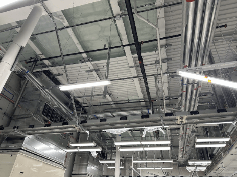 A ceiling view of an industrial building showing exposed pipes, ductwork, and fluorescent lighting fixtures.