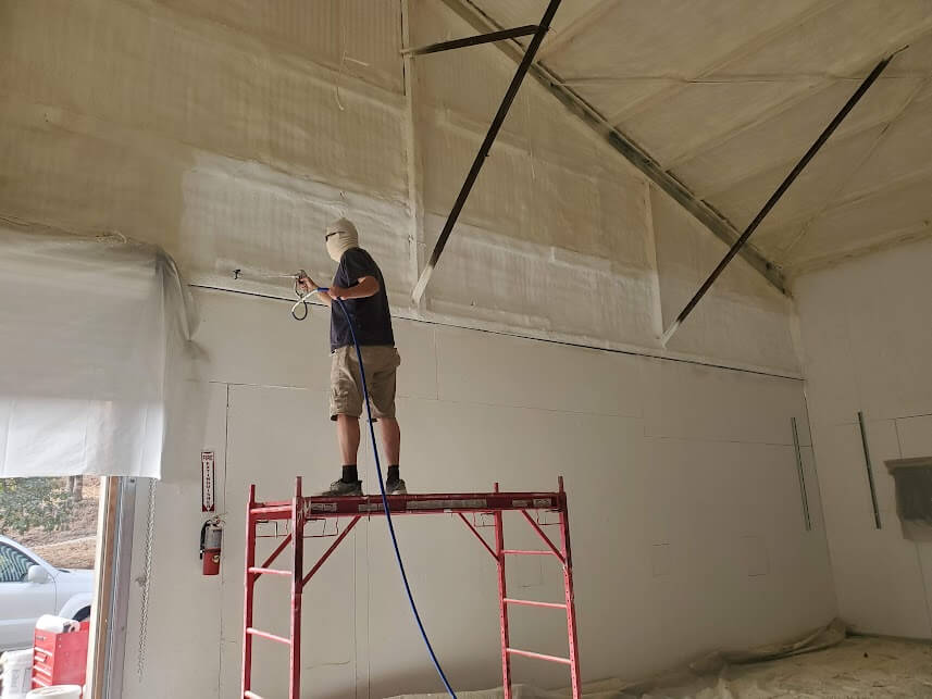 Worker applying spray foam insulation on an interior wall from scaffolding.