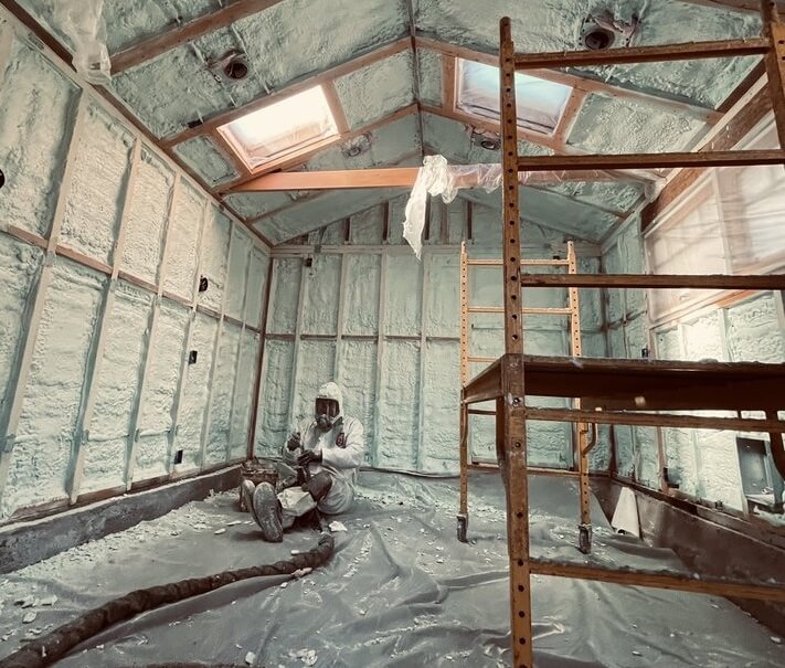 Worker in protective gear applying spray foam insulation in a room with unfinished walls and skylights.