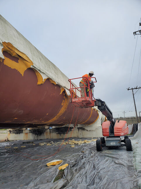 A worker in safety gear applying spray polyurethane foam insulation to a storage tank